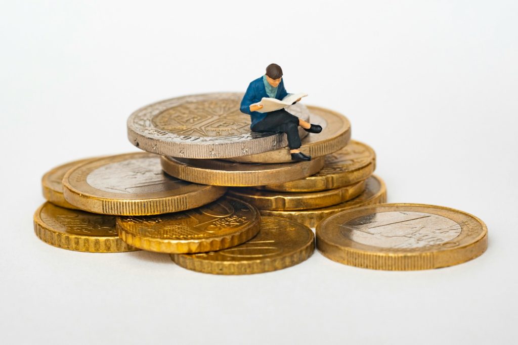 Miniature person sitting on stack of coins reading newspaper
