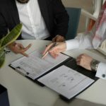 two men sitting at a table with papers and a pen